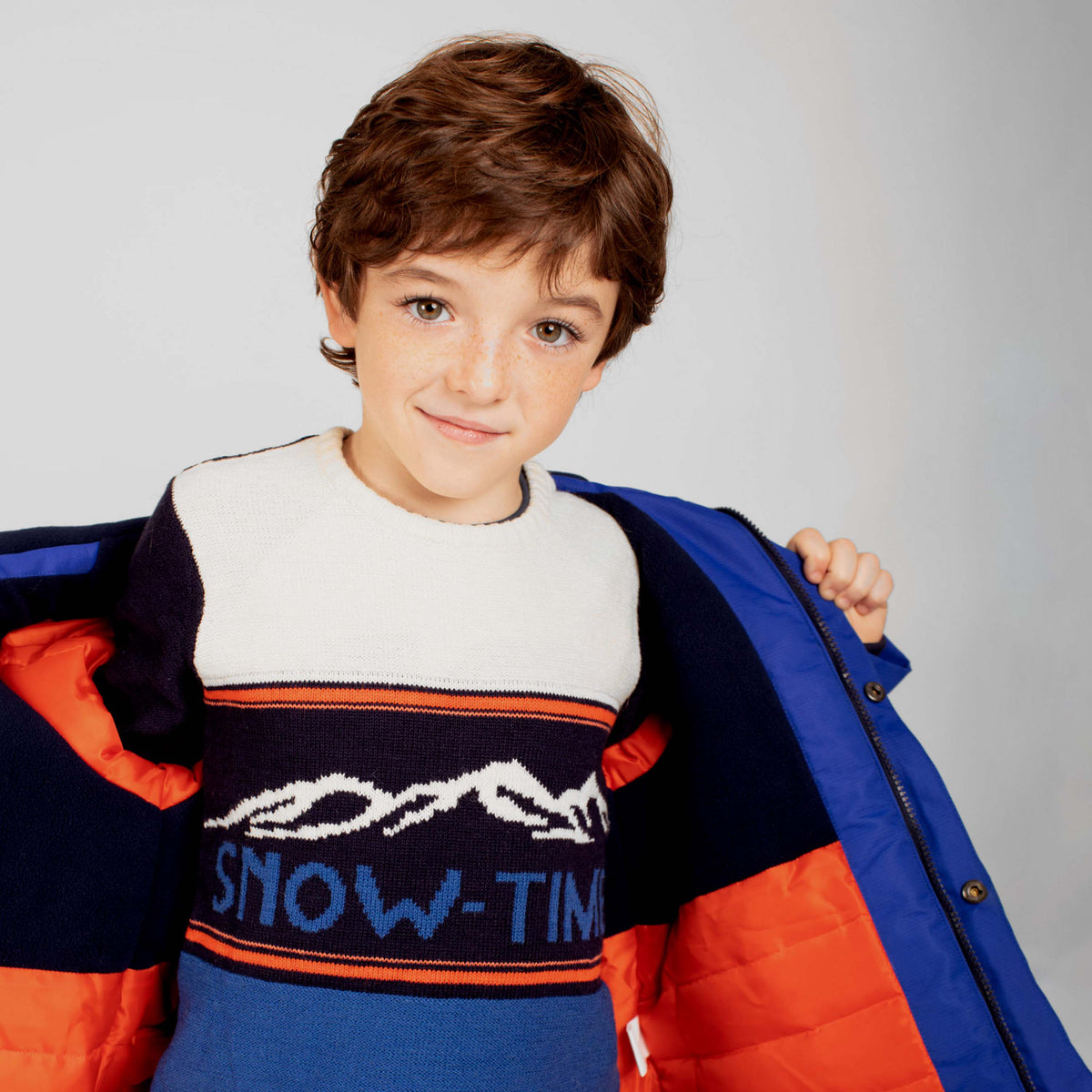 Niño sonriendo con jersey azul SNOW-TIME estampado de montañas y texto, foto lifestyle de medio cuerpo con abrigo abierto Charanga